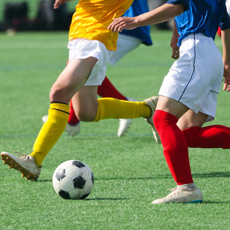 Two soccer players in action on a field; one player wears a yellow uniform with yellow socks, while the other wears a blue shirt, white shorts, and red socks. A black and white soccer ball is visible near their feet.