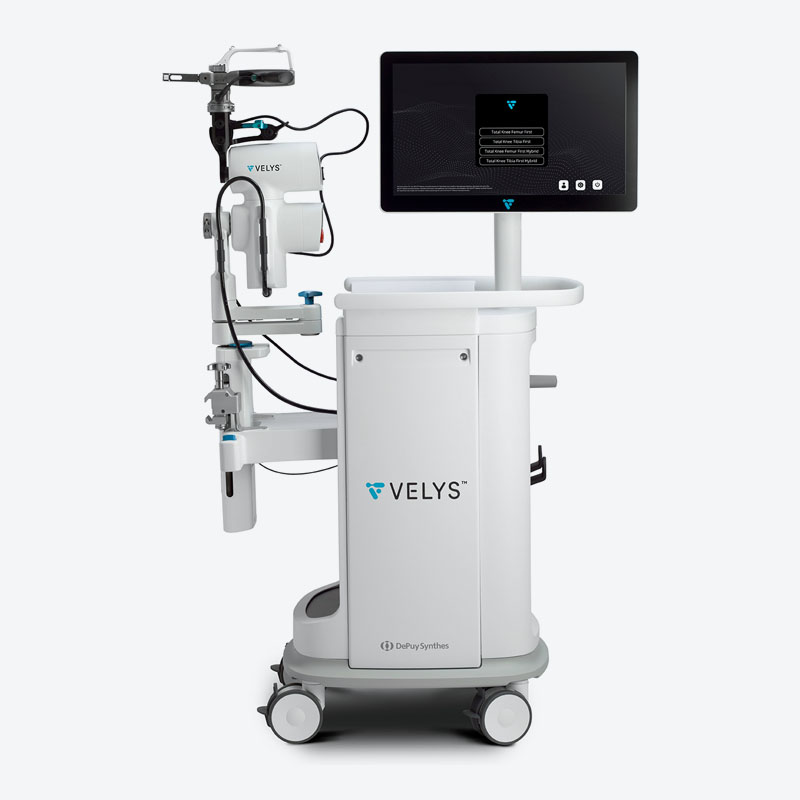 A white VELYS medical robotic system on wheels with an attached monitor, control panel, and mechanical arm, designed for surgical procedures. The DePuy Synthes logo is visible on the front.