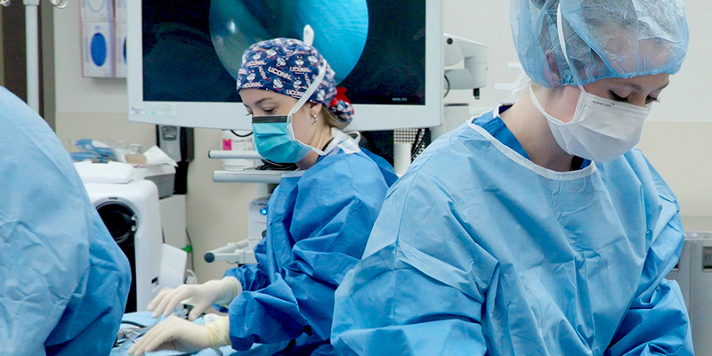Two medical professionals in blue surgical gowns, masks, and caps prepare instruments in an operating room. One arranges tools while another works at a table; a medical monitor displays an image in the background.
