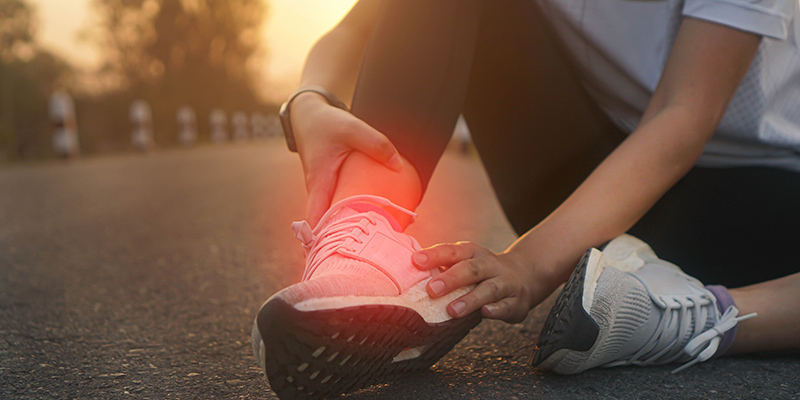 A person sitting on a road, holding their ankle in pain with a red glow highlighting the injured area, suggesting an ankle injury while wearing athletic shoes.