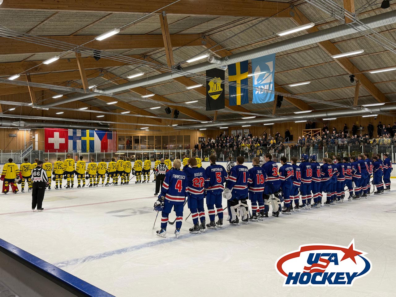 Two hockey teams, one in yellow and one in blue, line up on the ice facing flags, including Swiss, Swedish, and American, in a rink with spectators. The USA Hockey logo is on the ice in the foreground.