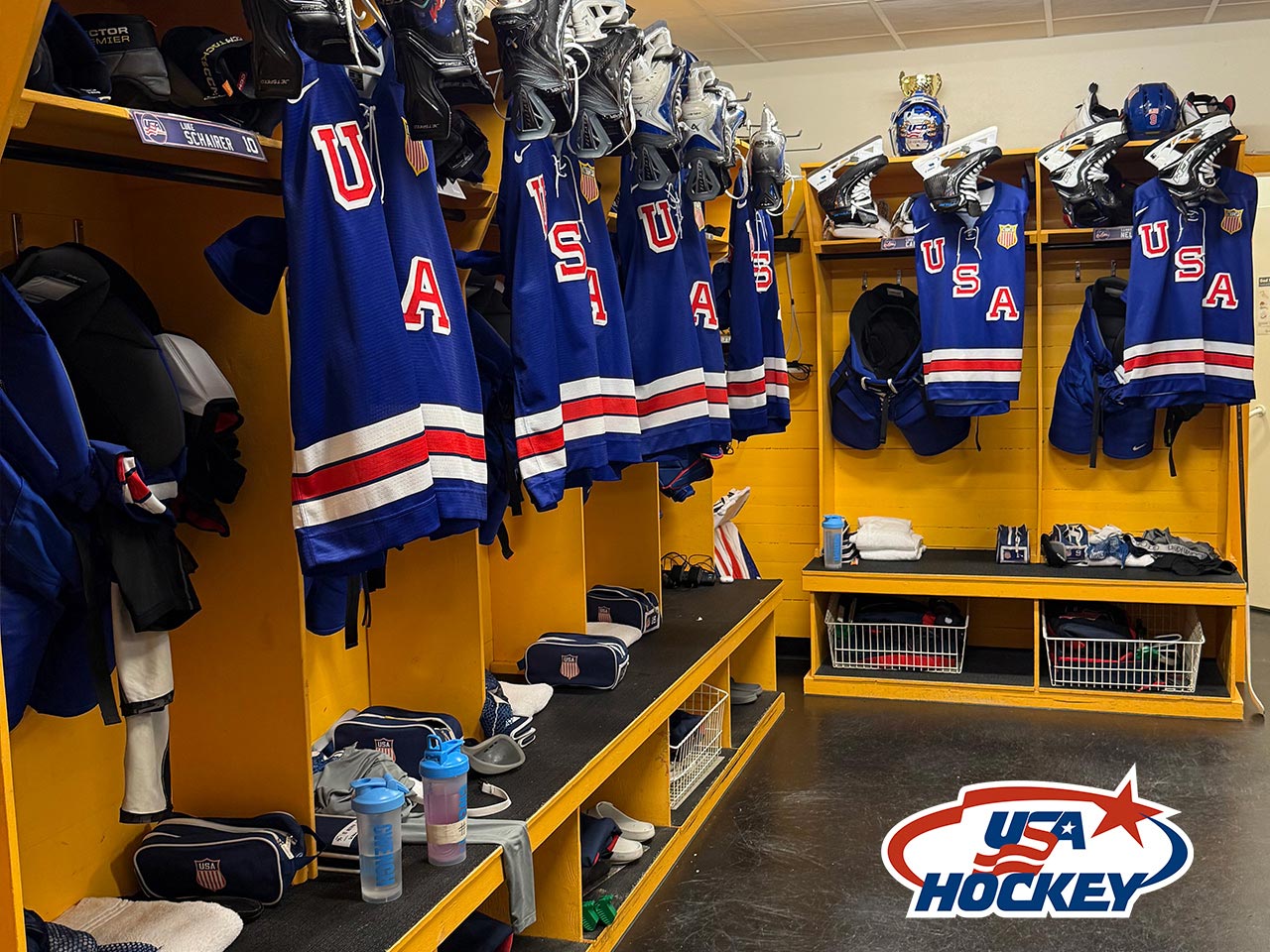 A locker room with blue USA hockey jerseys hanging in open lockers, equipment below, and the "USA Hockey" logo in the bottom right corner. Helmets and pads are organized neatly on shelves.