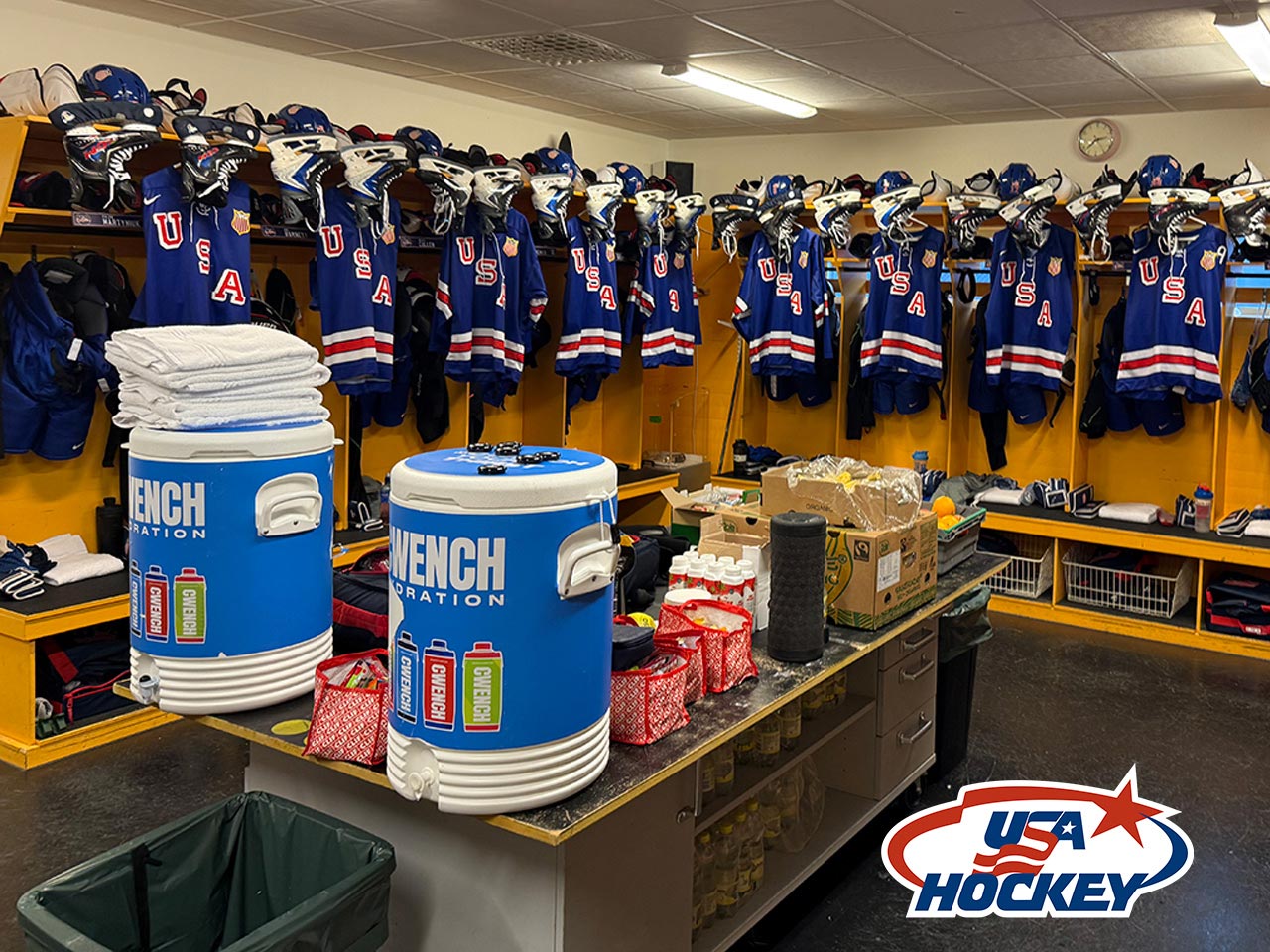A hockey locker room with blue "USA" jerseys hanging in cubbies, helmets and gear above. Two blue water coolers, towels, and supplies are on a table. The USA Hockey logo is in the bottom right corner.