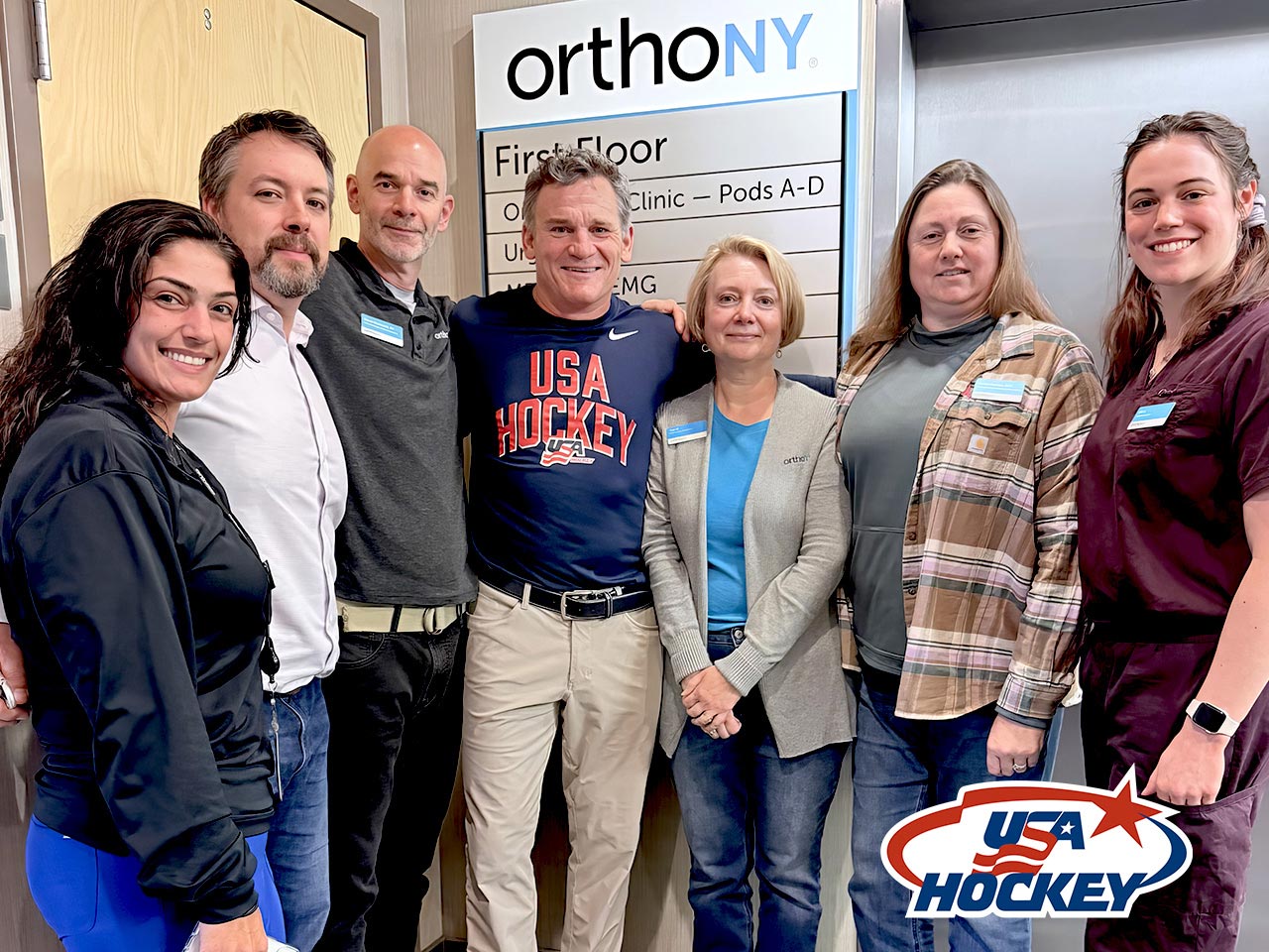 Seven people stand together and smile in front of an OrthoNY sign. One person wears a USA Hockey shirt. The USA Hockey logo is visible in the bottom right corner of the image.