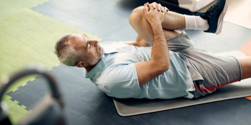 A middle-aged man with gray hair lying on an exercise mat indoors, stretching one leg toward his chest while keeping the other leg bent on the floor. He is wearing athletic shorts, a light shirt, and sneakers.