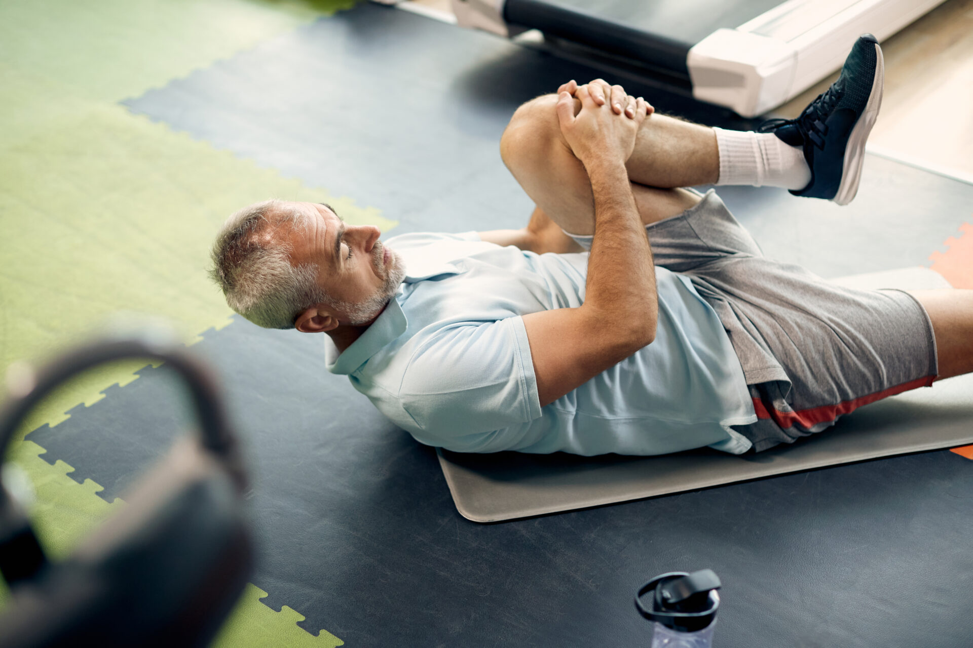 An older man in athletic wear lies on a mat at the gym, stretching by pulling one knee toward his chest. Exercise equipment and a water bottle are visible nearby.