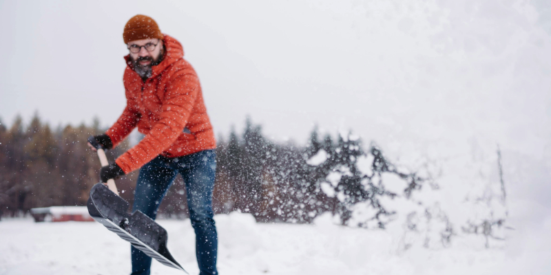 Man wearing an orange winter jacket shovels heavy snow outdoors, highlighting safety tips for shoveling snow and proper technique during cold winter weather.