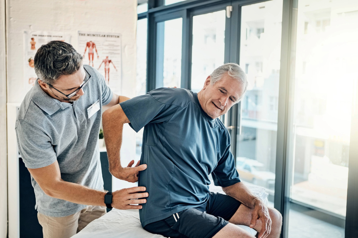 A physical therapist examines an older man’s lower back as he sits on an exam table, both indoors with large windows and medical posters in the background. The man appears to be in mild discomfort.