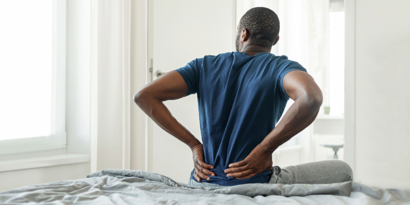 A man sitting on his bed holds onto his back, due to ongoing back pain.