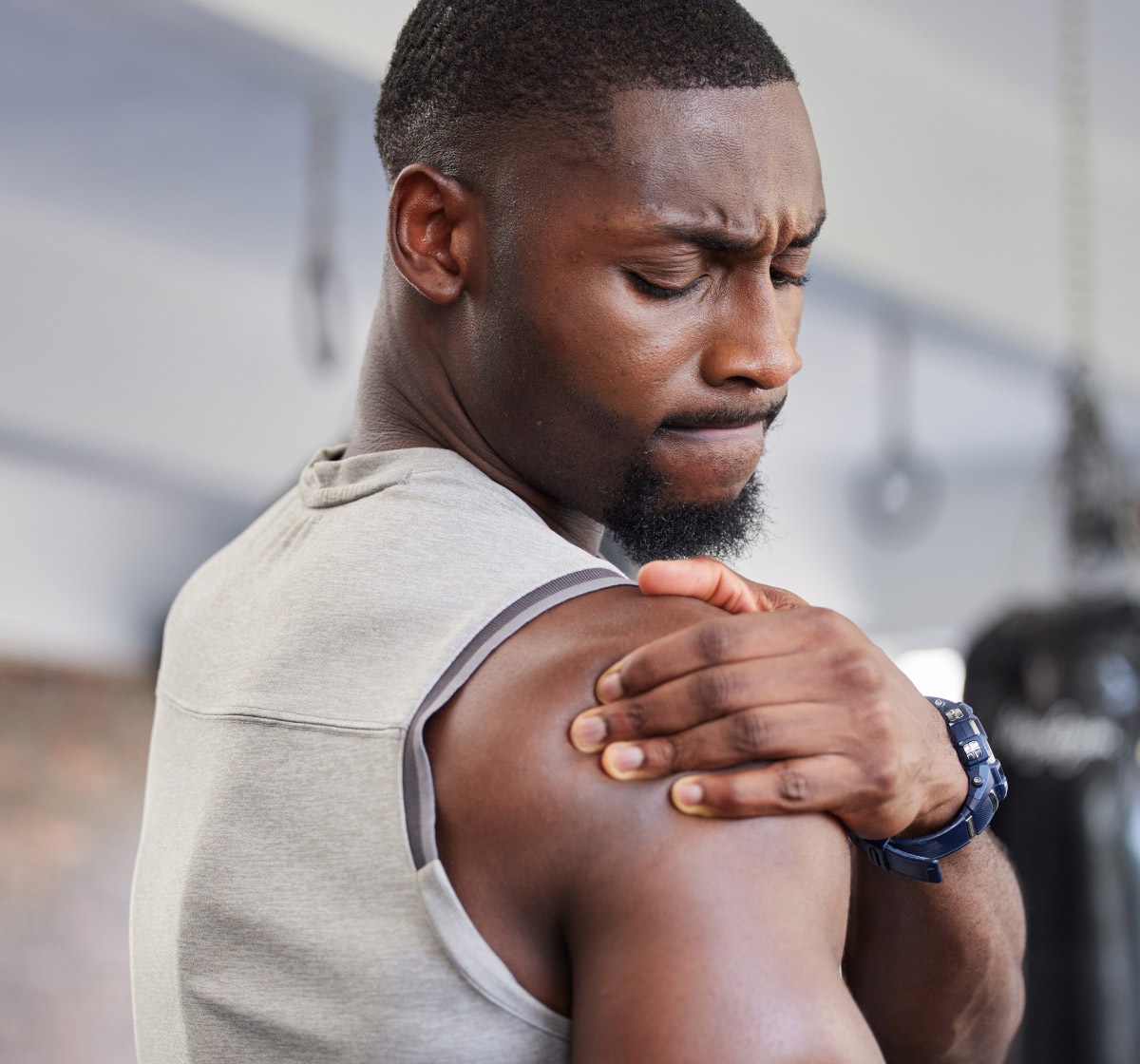 A man in a sleeveless shirt stands indoors, looking at and touching his sore shoulder with a pained expression on his face.