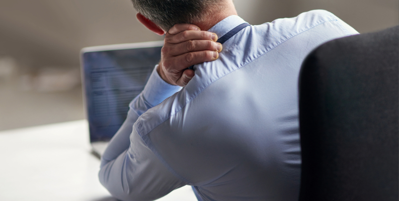 A man in a light blue shirt sits at a desk with a laptop, holding the back of his neck, appearing to experience discomfort or pain while working.