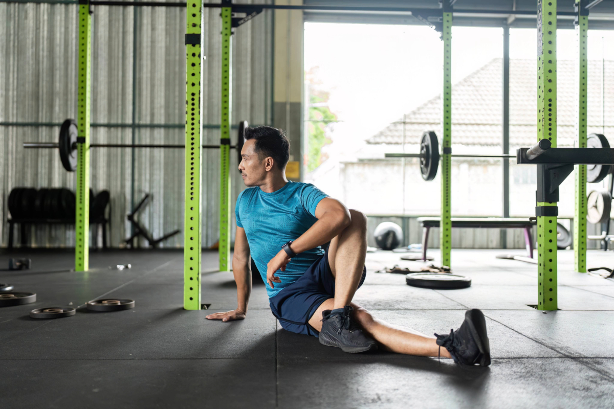A man in athletic wear stretches on the floor of a gym, performing a seated lower back rotational stretch with one leg bent to relieve lower back pain.