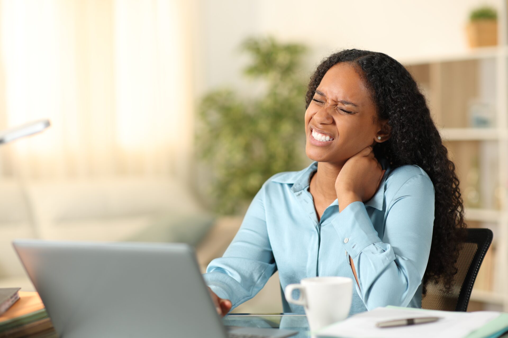 A woman sitting at a desk with a laptop, holding her neck and wincing in pain, suggesting discomfort or neck strain. A coffee cup and papers are on the desk, and the room appears bright and homey.