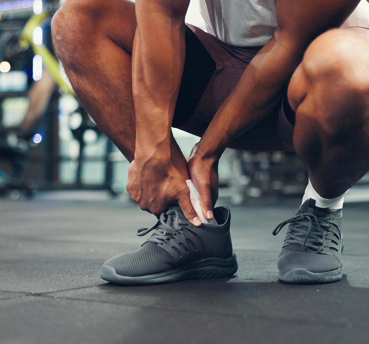 A person in athletic wear crouches on a gym floor, tying the laces of their gray running shoes. Their muscular arms and legs are visible, with gym equipment blurred in the background.