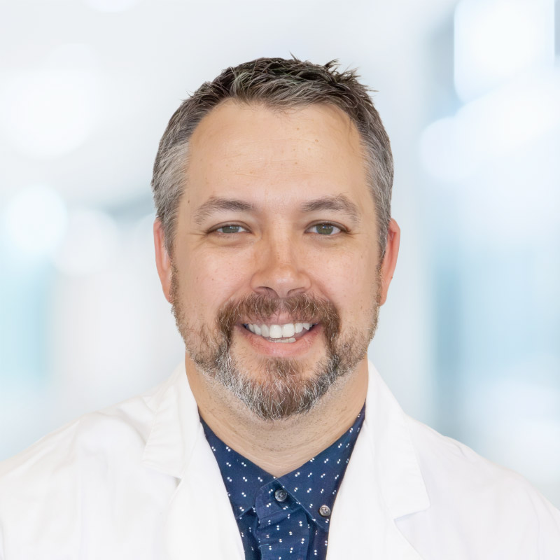 A smiling man with short brown and gray hair and a beard, wearing a white lab coat over a navy blue shirt with small white patterns, standing in a brightly lit, blurred indoor setting.