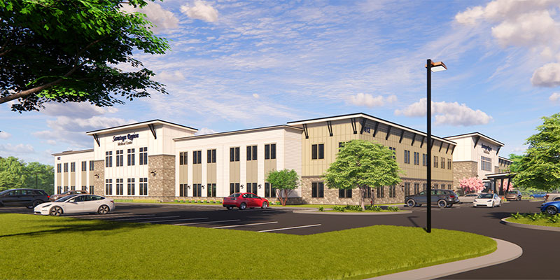 Modern, two-story building with large windows and stone accents, surrounded by trees and landscaping. Several cars are parked in a lot in front, with a clear blue sky and scattered clouds overhead.